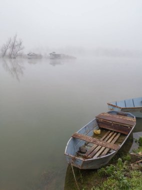 Sakin nehrin kıyısındaki eski paslı balıkçı tekneleri ve sislerin derinliklerindeki nehir adasının önündeki balıkçı kulübeleri. Kasvetli sonbahar manzarası kalın bir sisle kaplanmış..