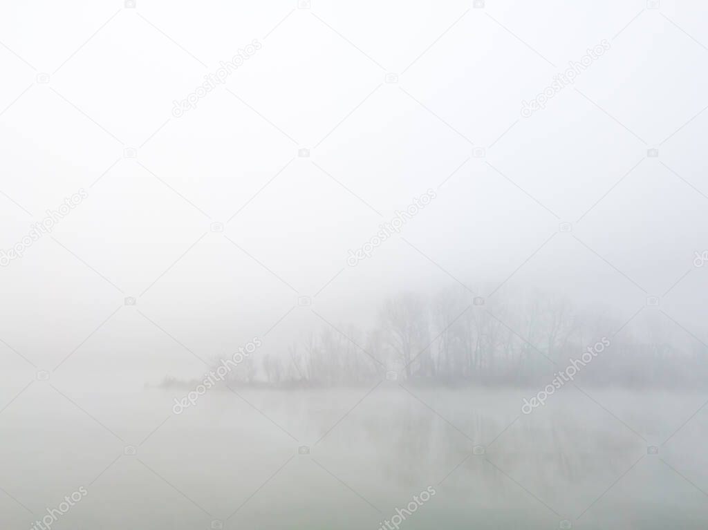 Peaceful autumn scene of mystical river island with a deciduous grove in deep fog early in morning. Calm and silent nature scene covered with thick fog.