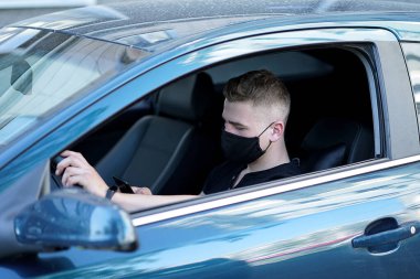 Adam direksiyonun arkasında siyah bir maskeyle oturuyor, siyah tişörtlü, eli direksiyonda, elinde, telefona bakıyor, yan fotoğrafa bakıyor.