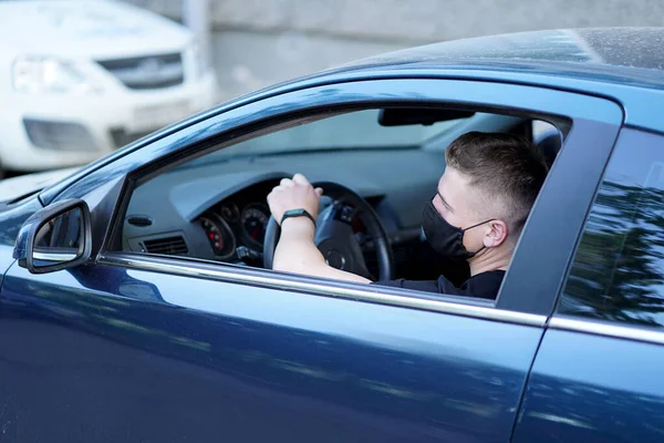 the guy is sitting in the car behind the wheel in a black mask, in a black t-shirt, hand on the steering wheel, watch on hand, looking out the open window, side photo