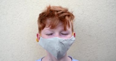 Koronavirüs salgını sırasında koruyucu maske takan küçük kızıl saçlı, mavi gözlü çocuk, kamera için poz veren,