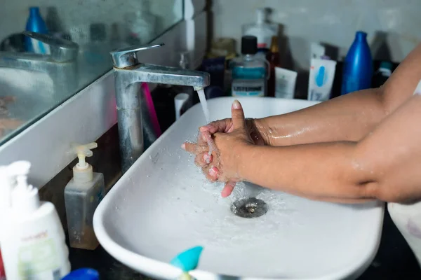 Female adult washing hands and arms properly with soap under the sink / wash basin in the washroom to battle Coronavirus. Bathroom console, hand wash with lot of products in the backdrop, space to add text