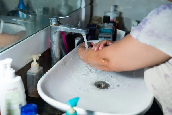 Female adult washing hands and arms properly with soap under the sink / wash basin in the washroom to battle Coronavirus. Bathroom console, hand wash with lot of products in the backdrop, space to add text