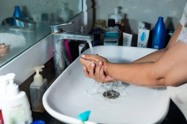 Female adult washing hands and arms properly with soap under the sink / wash basin in the washroom to battle Coronavirus. Bathroom console, hand wash with lot of products in the backdrop, space to add text