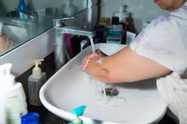 Female adult washing hands and arms properly with soap under the sink / wash basin in the washroom to battle Coronavirus. Bathroom console, hand wash with lot of products in the backdrop, space to add text