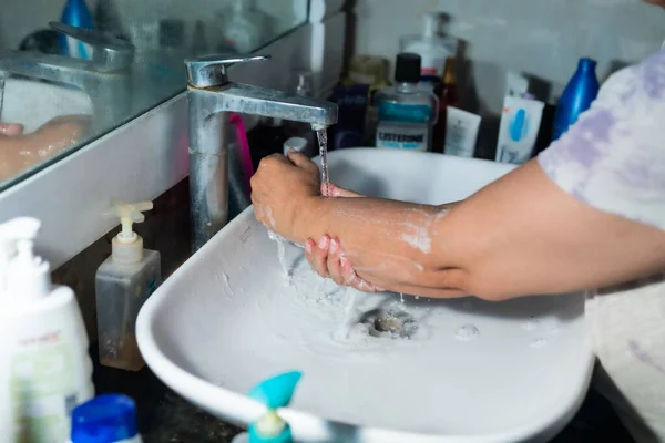 Female adult washing hands and arms properly with soap under the sink / wash basin in the washroom to battle Coronavirus. Bathroom console, hand wash with lot of products in the backdrop, space to add text