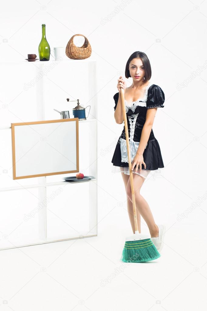 Jeune fille avec joli visage en costume de servante sexy maid noir belle posant avec balai de balayage vert près de vaisselle sur étagères isolé sur fond