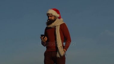 Noel Baba şapkası sakallı adam sigara içiyor ve Noel arifesinde akşam gökyüzü karşı onun smartphone cep telefonu ile bir sms yazıyor