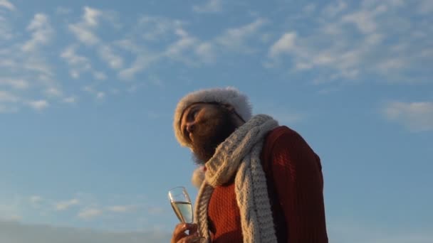 Junger bärtiger Weihnachtsmann mit Champagner spielt mit seiner Mütze — Stockvideo
