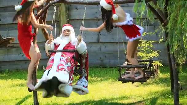 Santa Claus schwingt auf einer Holzschaukel mit zwei hübschen sexy Mädchen in weihnachtlichem, rotem Anzug und Hut — Stockvideo