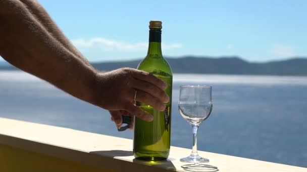 Чоловічі руки відкривають пляшку білого вина на балконі біля моря у відпустці, повільний рух — стокове відео