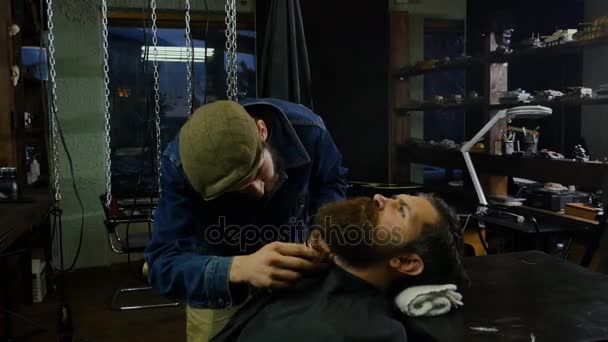Proceso de peinado. Peluquero corta la barba recortada de un joven barbudo en una elegante barbería moderna — Vídeos de Stock