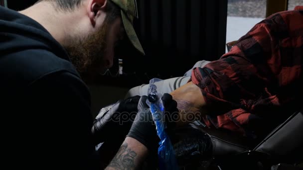 Tatuaggio artista barbuto dimostra da vicino il processo di ottenere tatuaggio nero con vernice. Master lavora in guanti sterili neri nel salone d'arte — Video Stock