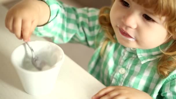 Niedliches Baby in grünem Hemd isst Milchdessert mit einem Löffel aus einem Spielzeugbecher — Stockvideo