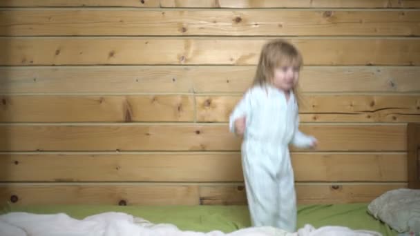 Cámara lenta de un niño pequeño y lindo o niño pequeño saltando felizmente en casa. El niño rizado en la cama salta en su pijama antes de irse a la cama. Niño es feliz y divertido en una casa de madera — Vídeo de stock