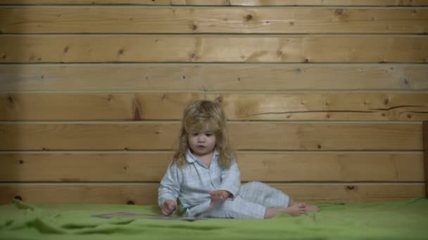 Χαριτωμένο μωρό με μακριά μαλλιά στις πυτζάμες διαβάζοντας ένα καλό βιβλίο πριν πάτε για ύπνο στο κρεβάτι. Ενδιαφέροντα των παιδιών εικονογραφημένο βιβλίο πριν πάτε για ύπνο — Αρχείο Βίντεο