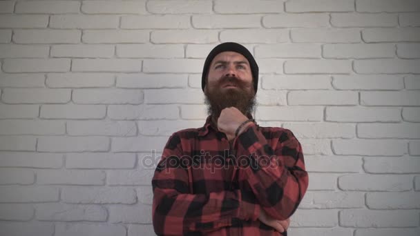 Un uomo premuroso con la barba che gli accarezza la barba, i baffi che volteggiano e gli incrocia le braccia sul petto. L'uomo barbuto pensa per strada vicino al muro di mattoni bianchi — Video Stock