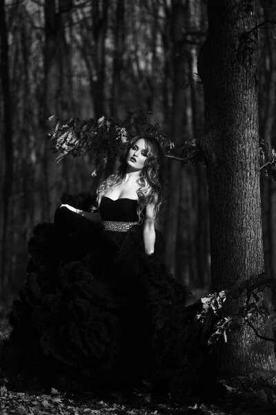 Young pretty sexy woman in elegant evening dress with skirt and long curly hair near tree in deep forest in sun light, black and white