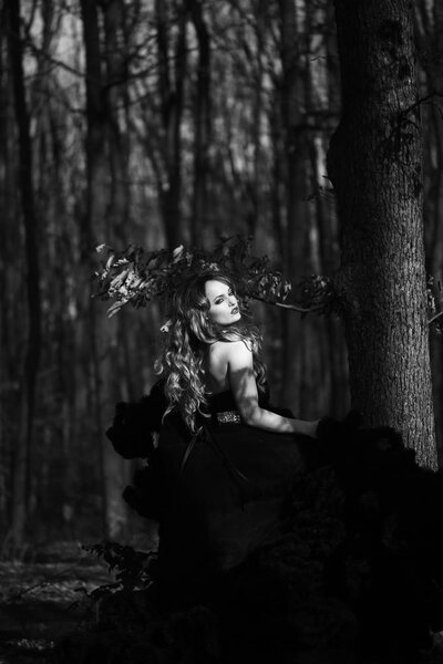Young pretty sexy woman in elegant evening dress with skirt and long curly hair near tree in deep forest in sun light, black and white