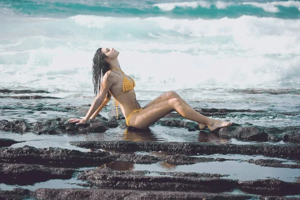 Hübsches Mädchen im sexy gelben Badeanzug sitzt am felsigen Strand — Stockfoto