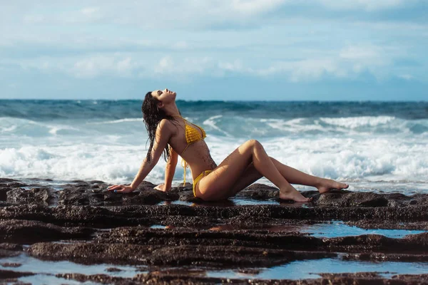 Красивая девушка в сексуальном жёлтом купальнике сидит на скалистом пляже — стоковое фото