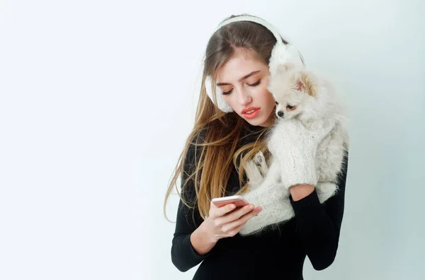 Jolie fille en cache-oreilles tenant un chien poméranien et utilisant un smartphone — Photo