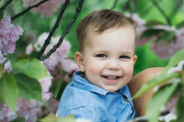 Schattige gelukkige jongen lachend in de armen van de moeder onder de bloeiende bloemen — Stockfoto