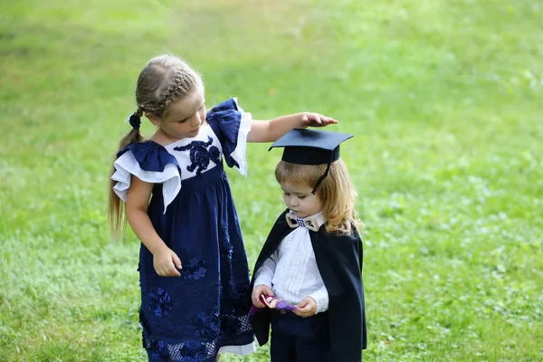 Милая девочка одевает маленького мальчика в шапку выпускника и халат — стоковое фото