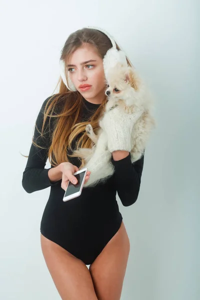 Красивая девушка в наушниках держит в руках померанскую собаку и пользуется смартфоном — стоковое фото