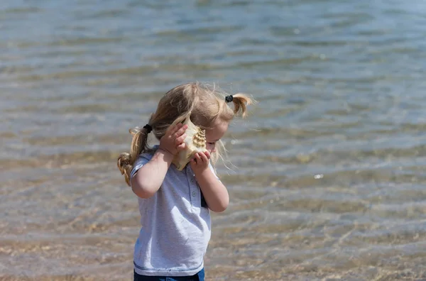 Cute baby boy listening to seashell on sea beach — Stock Photo, Image