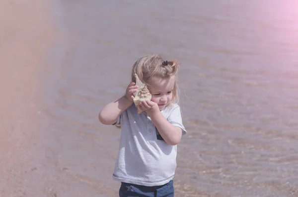 Lindo bebé niño escuchando concha marina en la playa del mar —  Fotos de Stock