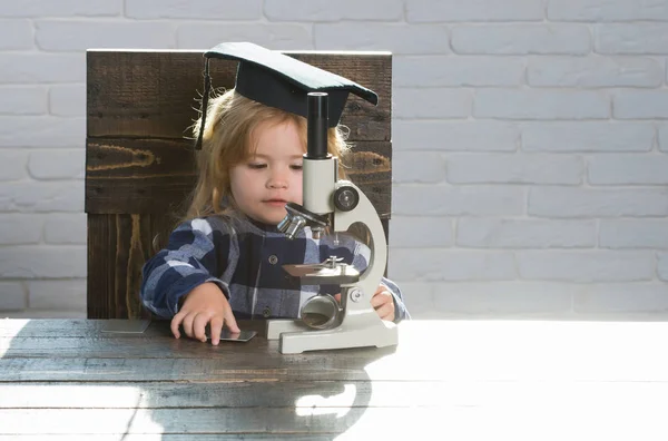 Criança inteligente em chapéu de graduação com estudo de microscópio no local de trabalho — Fotografia de Stock