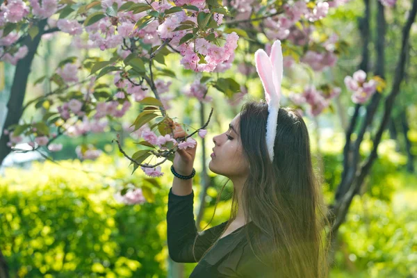 Χαριτωμένο κορίτσι με ρόδινα bunny αυτιά μυρίζοντας λουλούδια sakura — Φωτογραφία Αρχείου