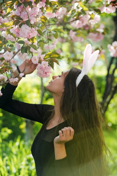 Meisje met roze bunny oren ruiken sakura bloemen in het voorjaar — Stockfoto