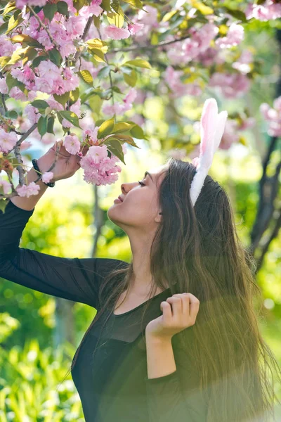 Meisje met roze bunny oren ruiken sakura bloemen uit boom — Stockfoto