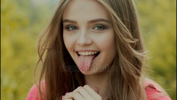 Linda chica muestra su lengua y mira a la cámara. Mujer joven se burla, coquetea. Ojos verdes, pelo largo. Chica en la naturaleza de primavera, el viento sopla su pelo. Atractiva mujer sexy. Retrato de modelo alegre — Vídeos de Stock