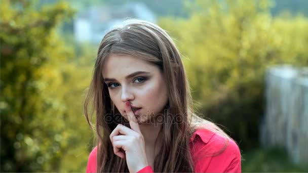 La chica sostiene el dedo cerca de sus labios. El acertijo de la joven. Chica joven en la naturaleza con el pelo largo. Emociones, coqueteo, boca sexy. Mujer con camiseta rosa, pidiendo silencio, silencio. Estudiante atractivo — Vídeos de Stock