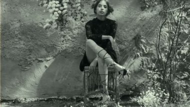 Saç, saç, retro makyaj, siyah ve beyaz video çiçekli güzel saç modeli ile oynarken eski duvara bahçesinde kız. Bayan bir hasır sandalye üzerinde ince ayaklı