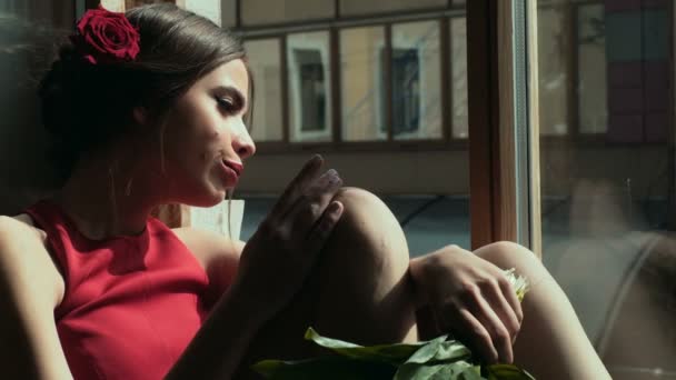 Красива дівчина з букетом конусів сидить на майданчику біля вікна і дивиться у вікно. Самотня дівчина з червоними квітами у волоссі, красивою зачіскою та макіяжем. Літній смуток — стокове відео