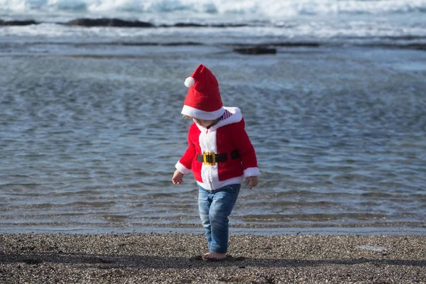 Kid in santa costume walking barefoot on sea beach — Stock Photo, Image