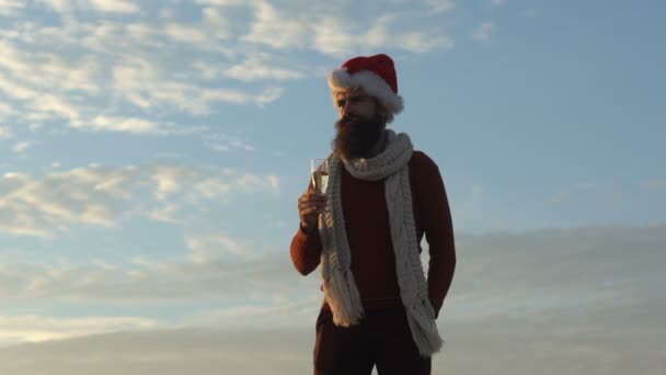 Santa Claus celebra con champán. Hombre barbudo en Santa Sombrero contra el cielo azul bebidas bebida, vino blanco. Vacaciones de Navidad e invierno, expectativa de Año Nuevo. Fiesta de Año Nuevo, soledad festiva — Vídeo de stock