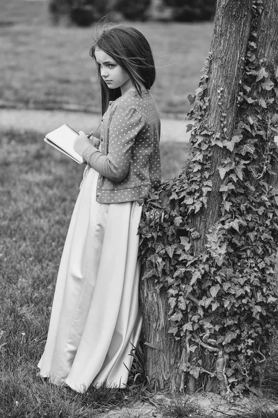 Dziewczyna w sukience z Notatnik i długopis, w pobliżu drzewa — Zdjęcie stockowe