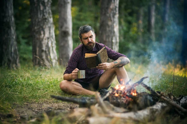 Хіпстерський піший турист з книгою і кухоль під багаттям у лісі — стокове фото
