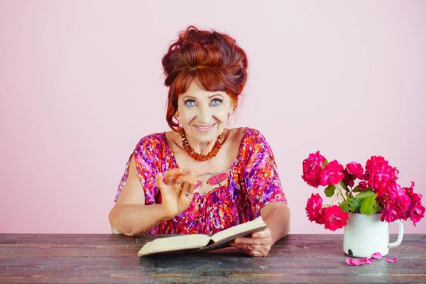 Oude vrouw lezen boek met glazen op bloemen. — Stockfoto