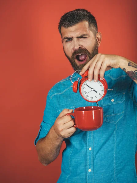 Hombre con barba en la cara somnolienta con taza, despertador —  Fotos de Stock