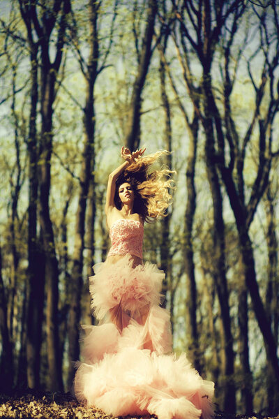 Beautiful young woman in pink glamour dress with long curly hair dancing in sunny forest