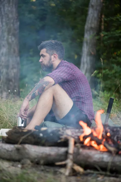 Хипстерский турист с кружкой отдыхает у костра в лесу — стоковое фото