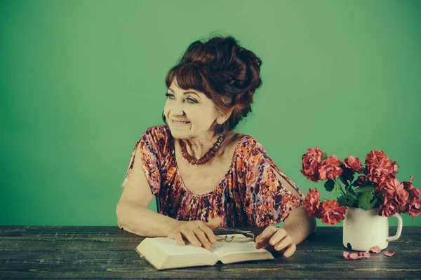 Oude vrouw lezen boek met glazen op bloemen. — Stockfoto