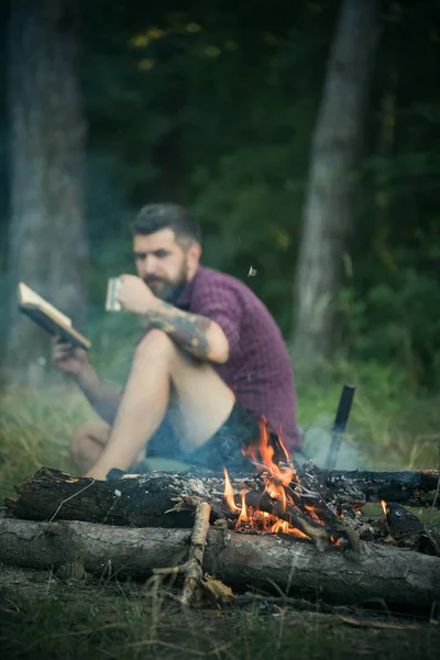 Hipster randonneur avec livre et tasse au feu de joie dans la forêt — Photo