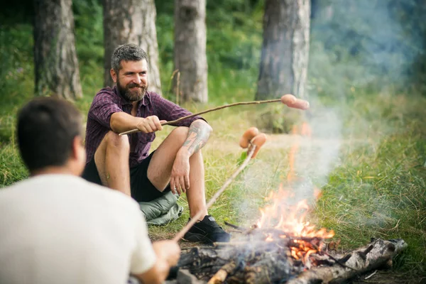 Друзі чоловіки смажать ковбаски на палицях на багатті в лісі — стокове фото